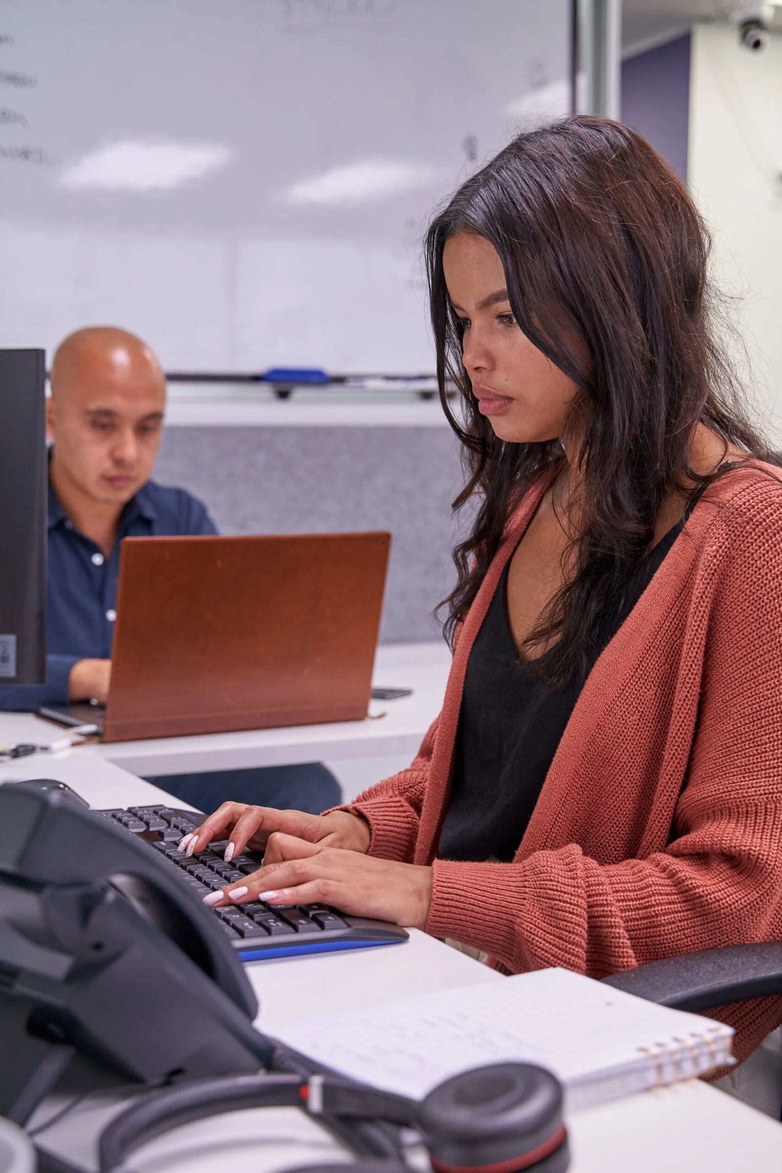 Woman in coral cardigan working at computer in office with man in background