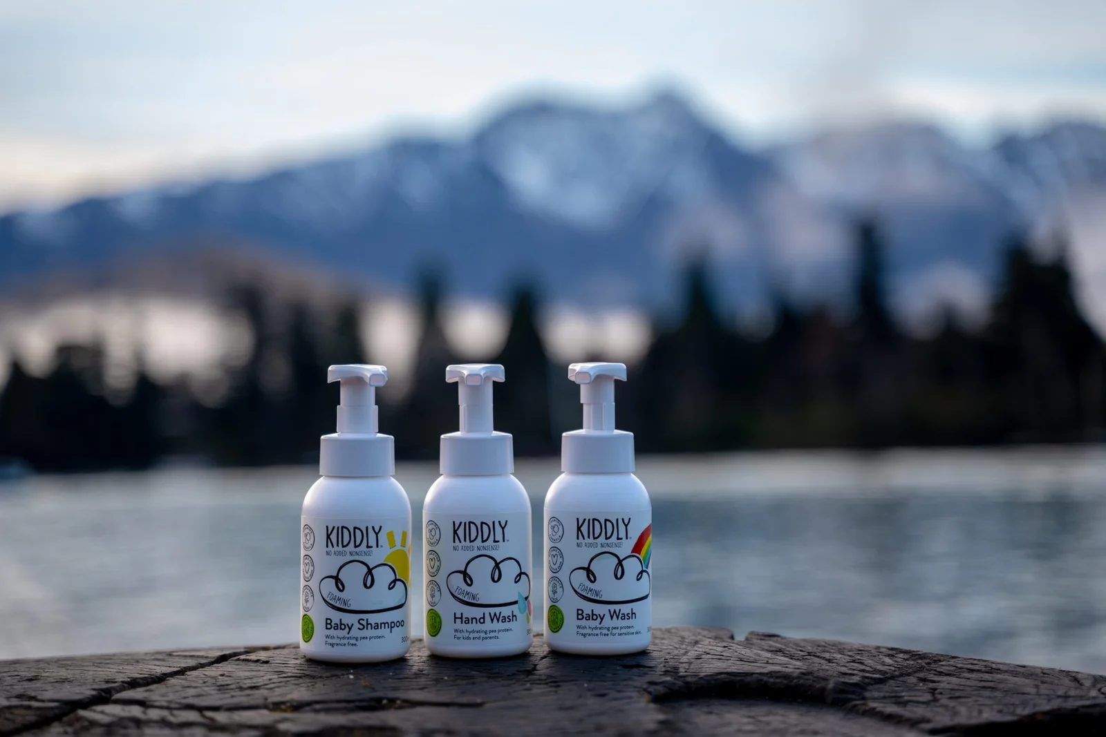 Three Kiddly brand baby product bottles on a wooden surface with a lake and mountains in the background