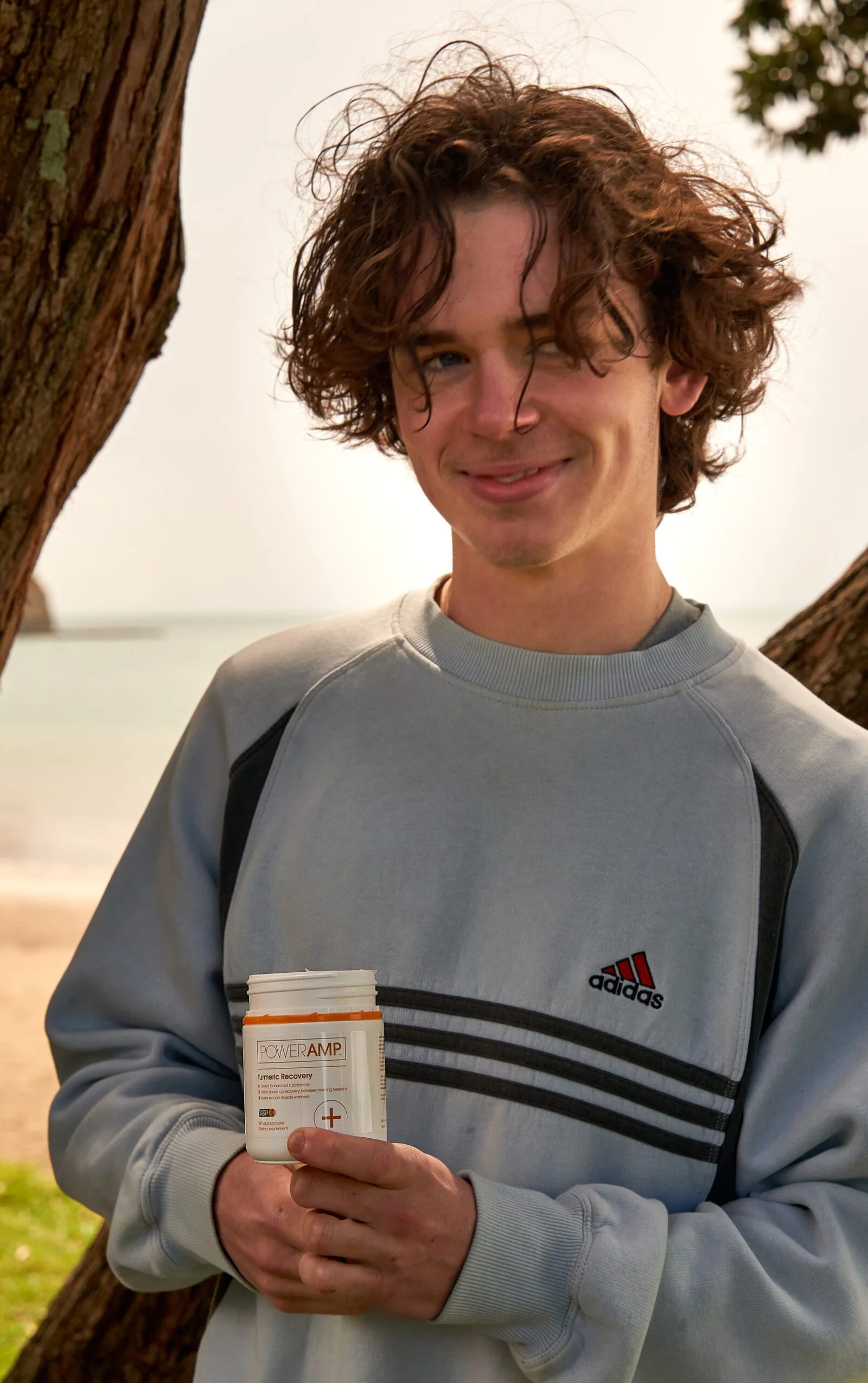 Young man with curly brown hair, wearing an Adidas sweatshirt, smiling and holding a white PowerAmp supplement bottle, with a beach and ocean in the background