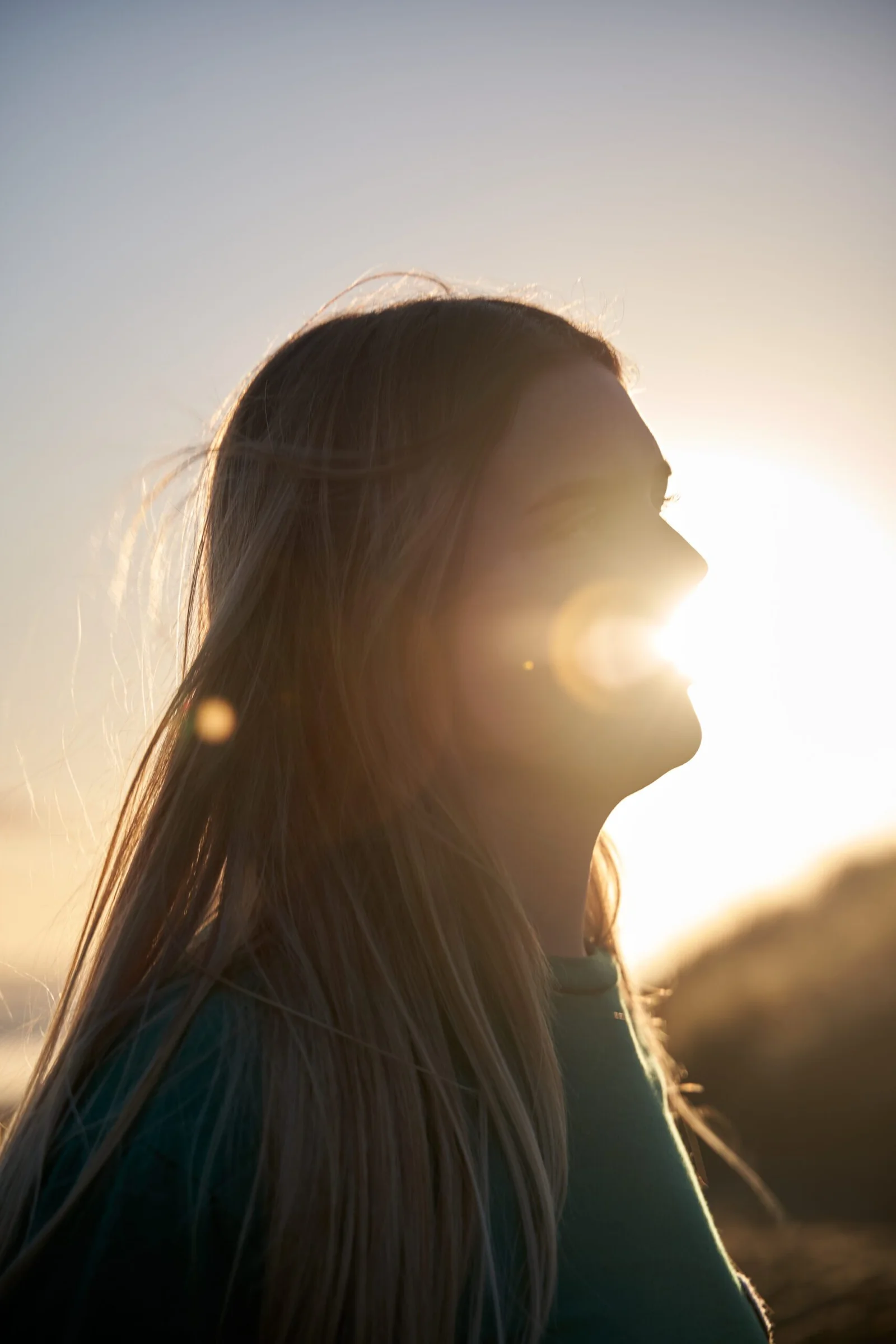 Side profile of a person with long blonde hair, silhouetted against a bright sunset or sunrise, with sun flare visible