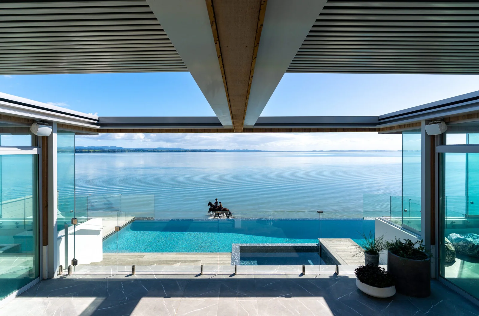 Luxurious outdoor living space with an infinity pool overlooking a vast lake or ocean, with a person on horseback in the distance