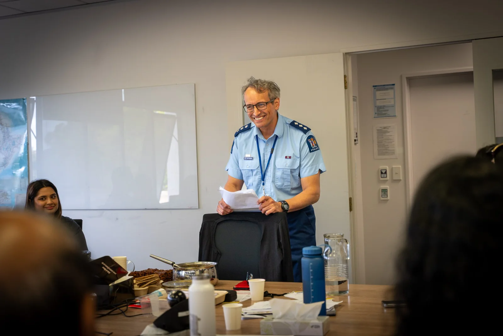Man in a police uniform smiling and holding papers, standing behind a chair at a conference table with other people in the foreground