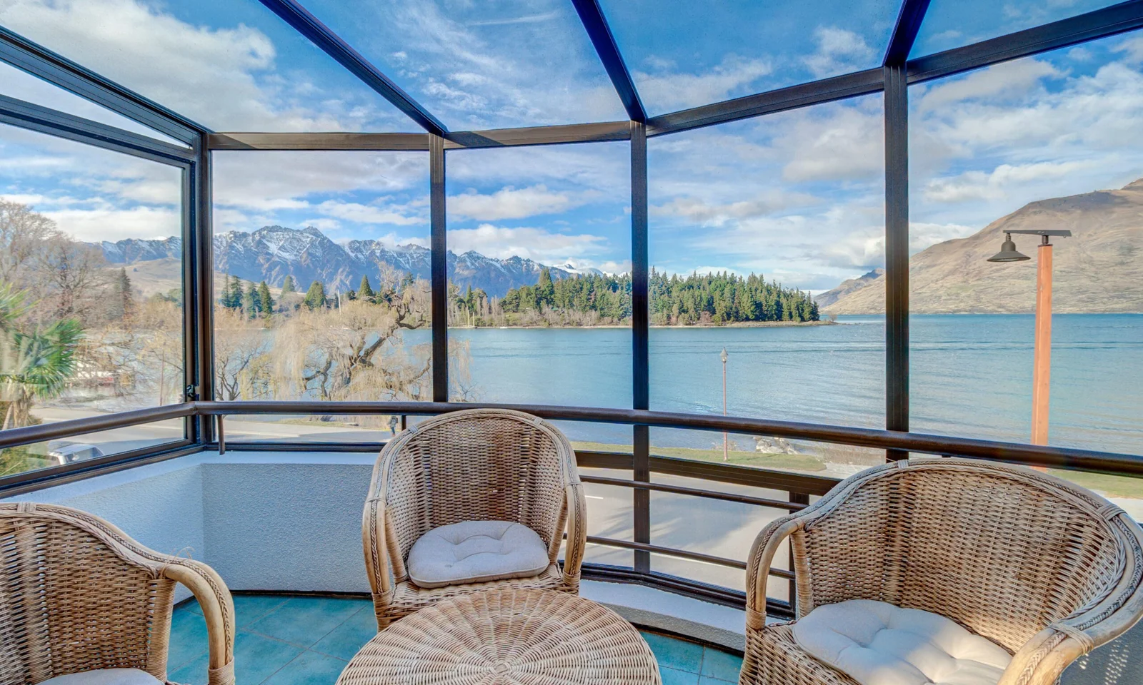 A sunroom or enclosed balcony with wicker furniture, offering a panoramic view of a lake and snow-capped mountains