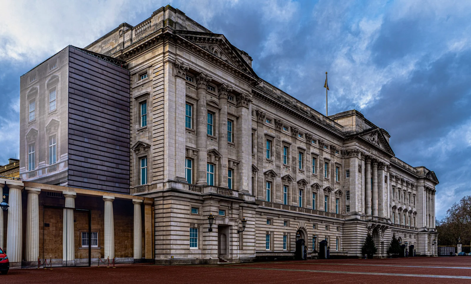Buckingham Palace under a dramatic cloudy sky, showing its classical architecture, columns, and numerous windows, with a large open paved area in the foreground