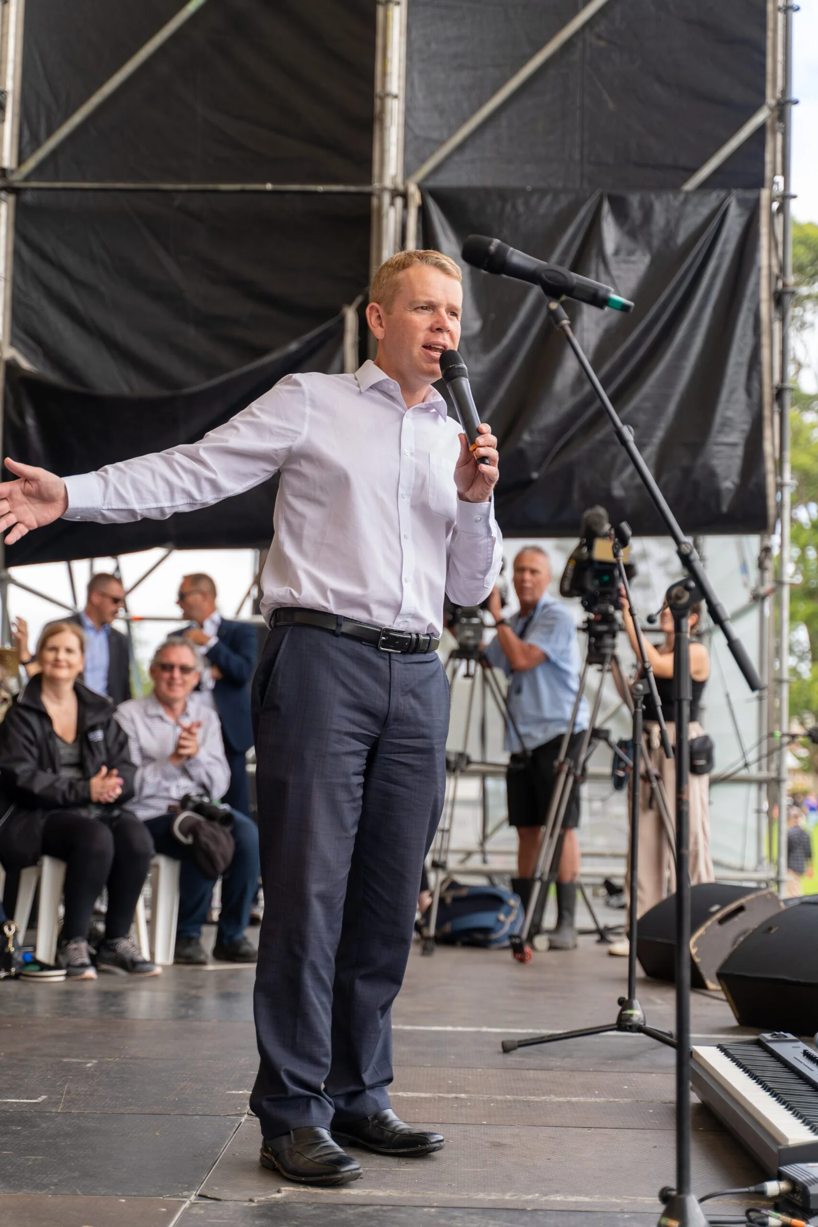 Man in white shirt speaking into microphone at outdoor event with audience and cameras