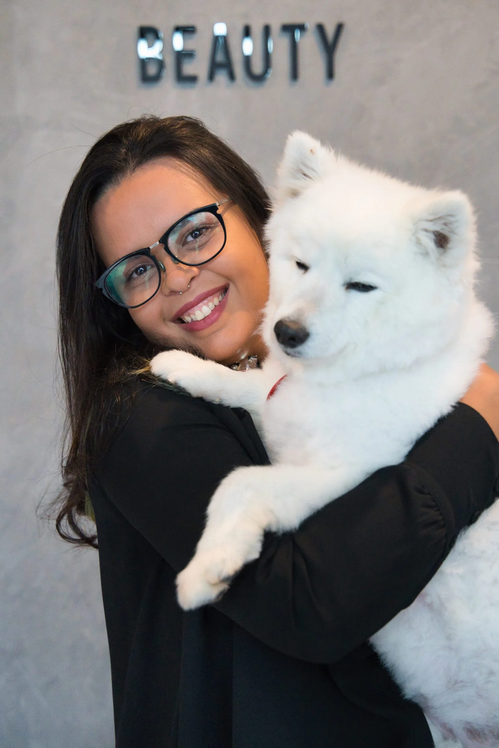 Woman with glasses holding white fluffy dog in front of beauty sign