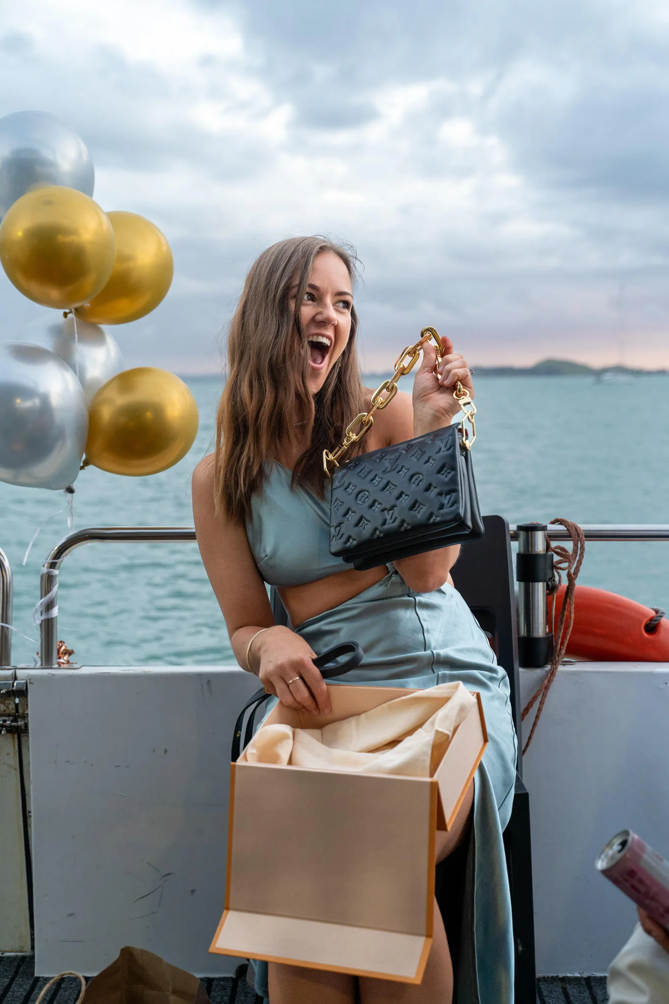Woman on a boat excitedly opening a gift box with a black handbag